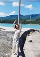 A woman in a white dress leaning against a pole by the water.